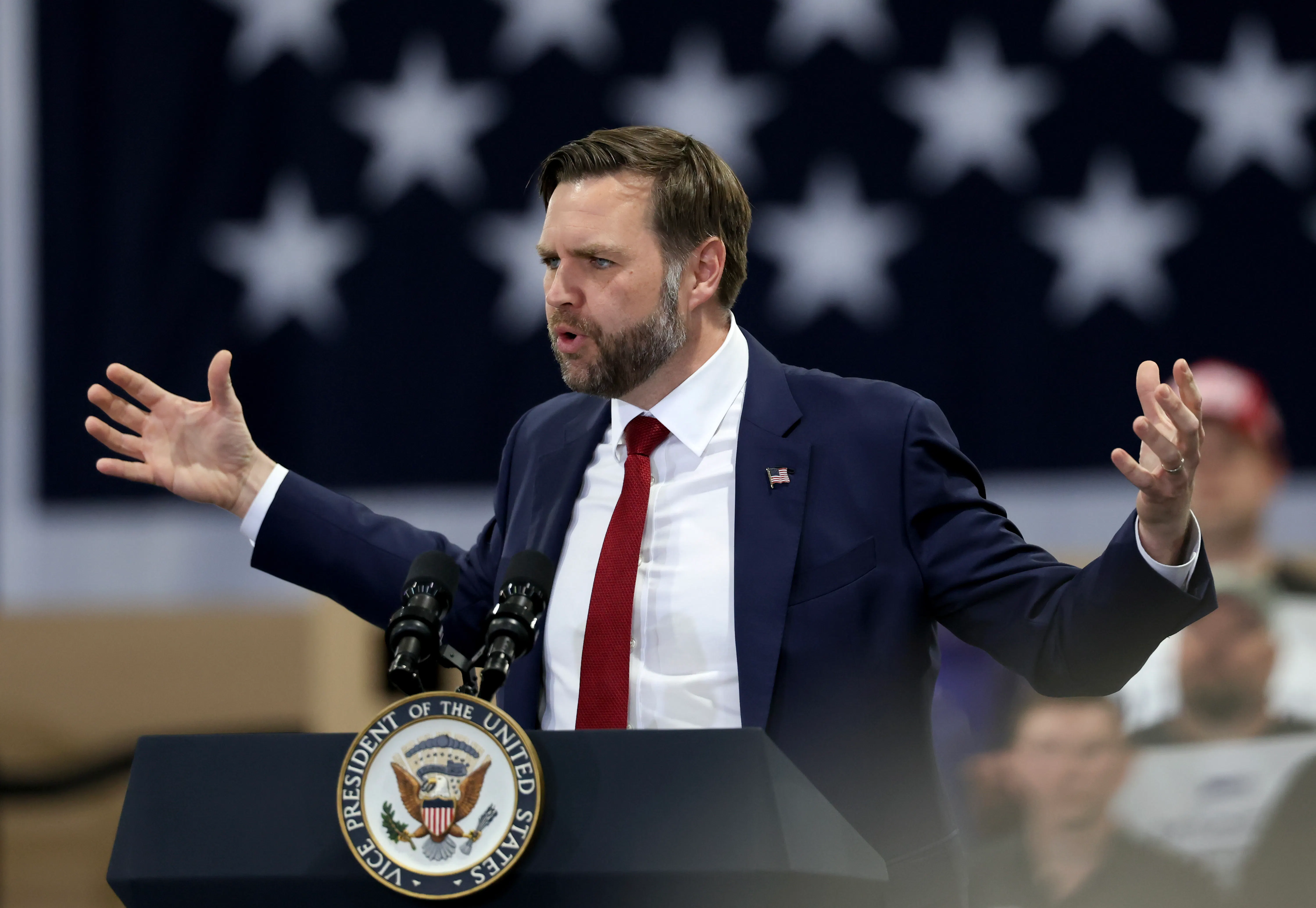 Vice President JD Vance holds his arms up and out to the side while speaking at a podium 