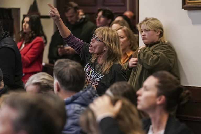 A woman in a crowd yells and points her finger at someone not pictured, as others listen.