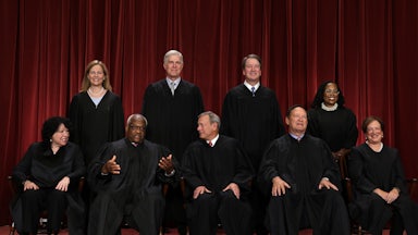 This image shows the nine justices against a red drapery background, with Sotomayor, Thomas, Roberts, Alito, and Kagan sitting, while the others stand behind.