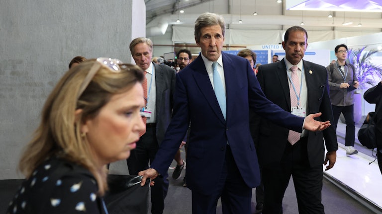John Kerry, flanked by others, holds his arm out in a questioning pose.
