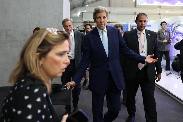 John Kerry, flanked by others, holds his arm out in a questioning pose.
