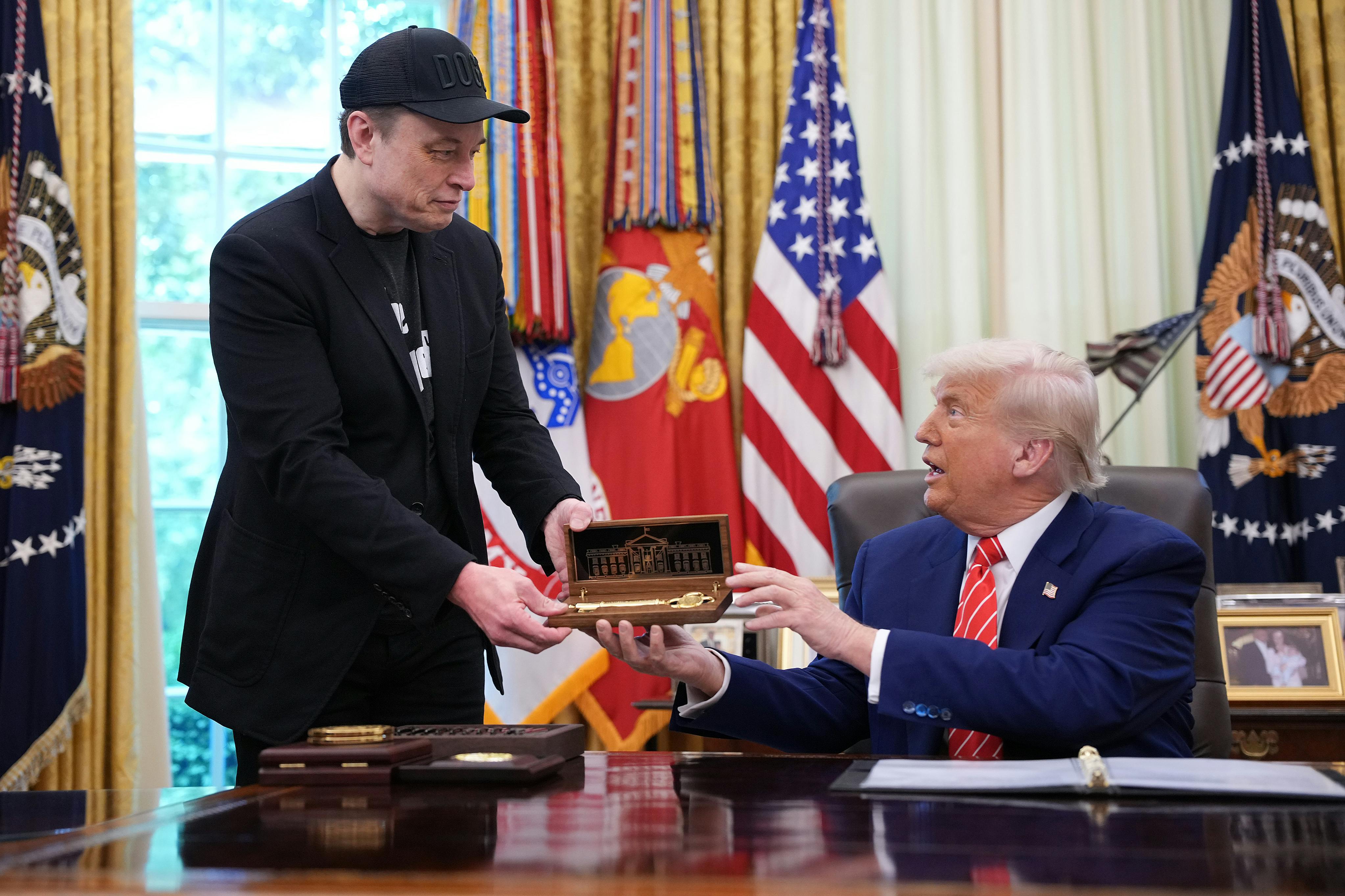 Donald Trump, seated behind his desk in the Oval Office, hands Elon Musk (standing above him) a golden key.