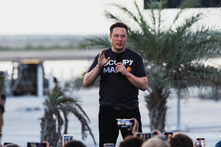 Elon Musk talks on stage holding a mic and wearing an "Occupy Mars" t-shirt
