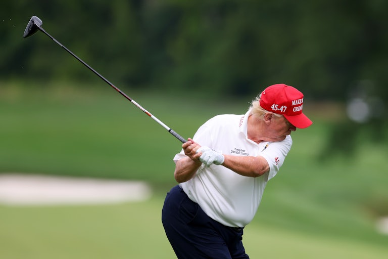 Donald Trump swings a golf club while wearing a red MAGA cap.