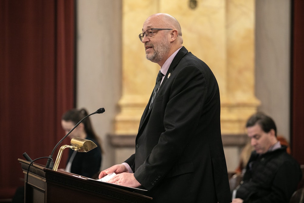 Gary Click stands at a lectern with other people seated in the background.