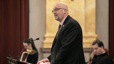 Gary Click stands at a lectern with other people seated in the background.