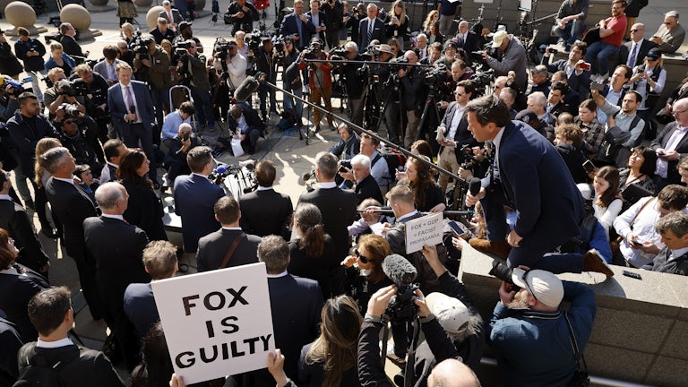 A crowd of people at a press conference. Someone in the background holds a sign that reads "Fox is guilty."