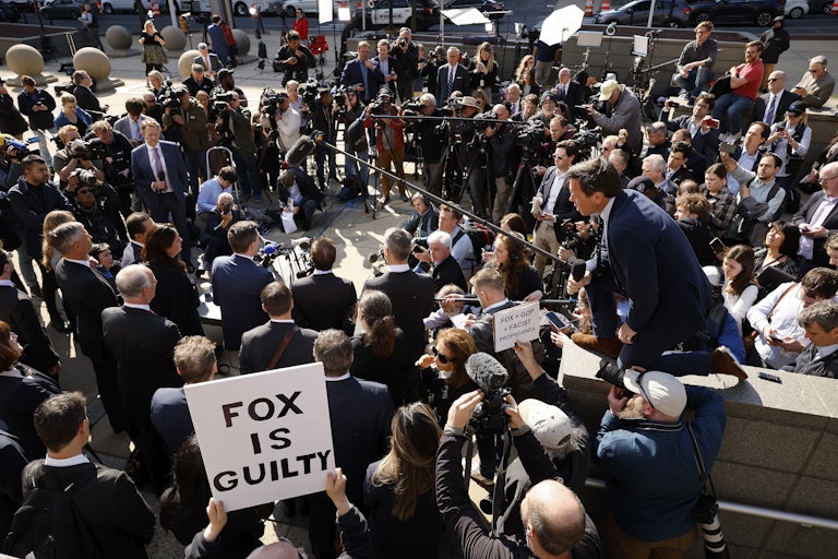 A crowd of people at a press conference. Someone in the background holds a sign that reads "Fox is guilty."