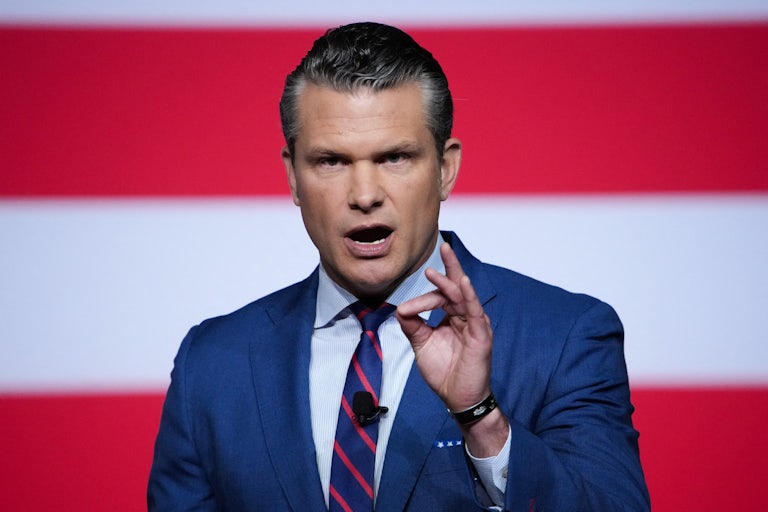 Pete Hegseth makes a hand gesture for emphasis while speaking in front of a U.S. flag.