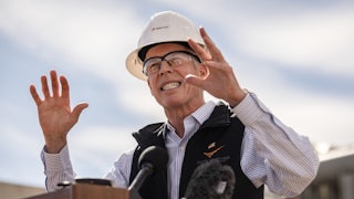 Energy Secretary Chris Wright wears a hard hat and eye protection glasses. He gestures with both hands while speaking at a podium.