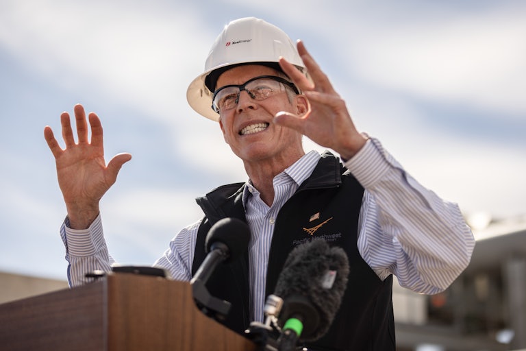 Energy Secretary Chris Wright wears a hard hat and eye protection glasses. He gestures with both hands while speaking at a podium.