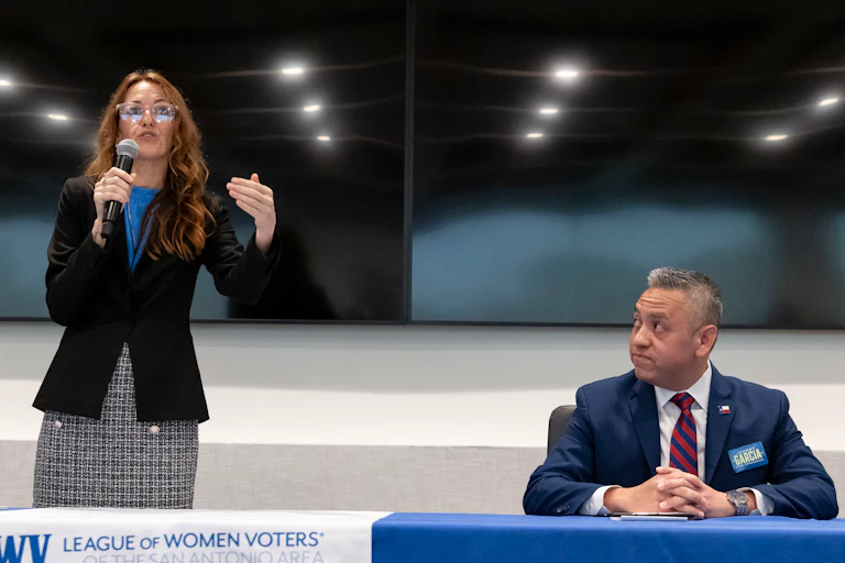 Democratic House primary candidate Maureen Galindo stands at a table. She holds a microphone and gestures while speaking. Primary candidate Johnny Garcia sits next to her.
