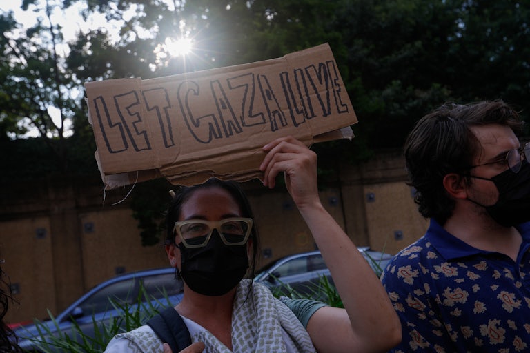 A protester wearing a keffiyeh and a face mask holds a carboard sign reading "Let Gaza Live."