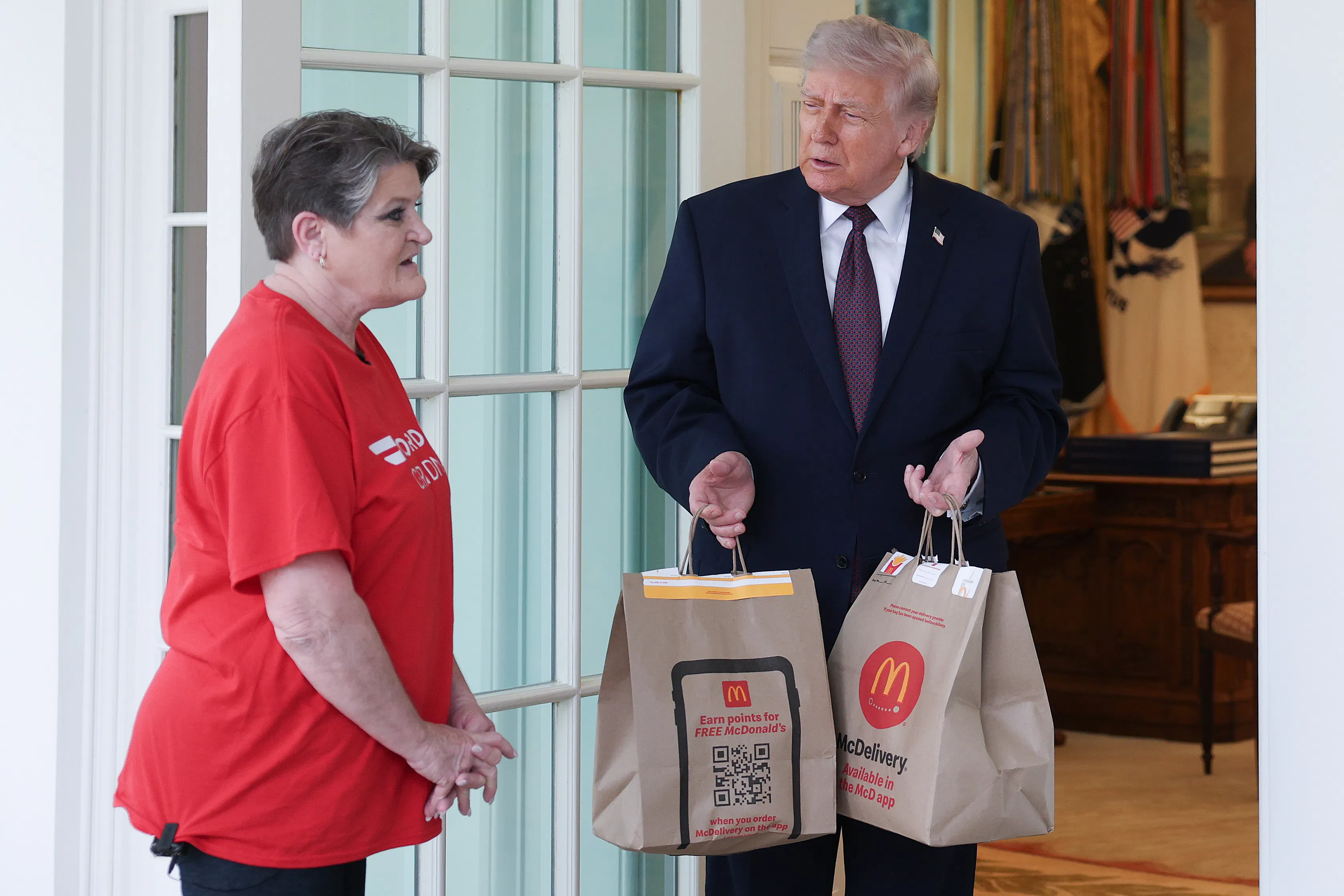 Donald Trump stands outside the Oval Office, holding McDonald's takeout bags while he watches DoorDash driver Sharon Simmons speak