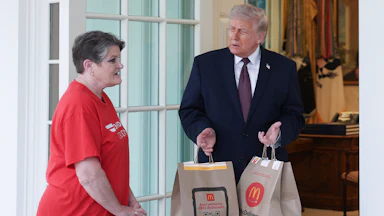 Donald Trump stands outside the Oval Office, holding McDonald's takeout bags while he watches DoorDash driver Sharon Simmons speak