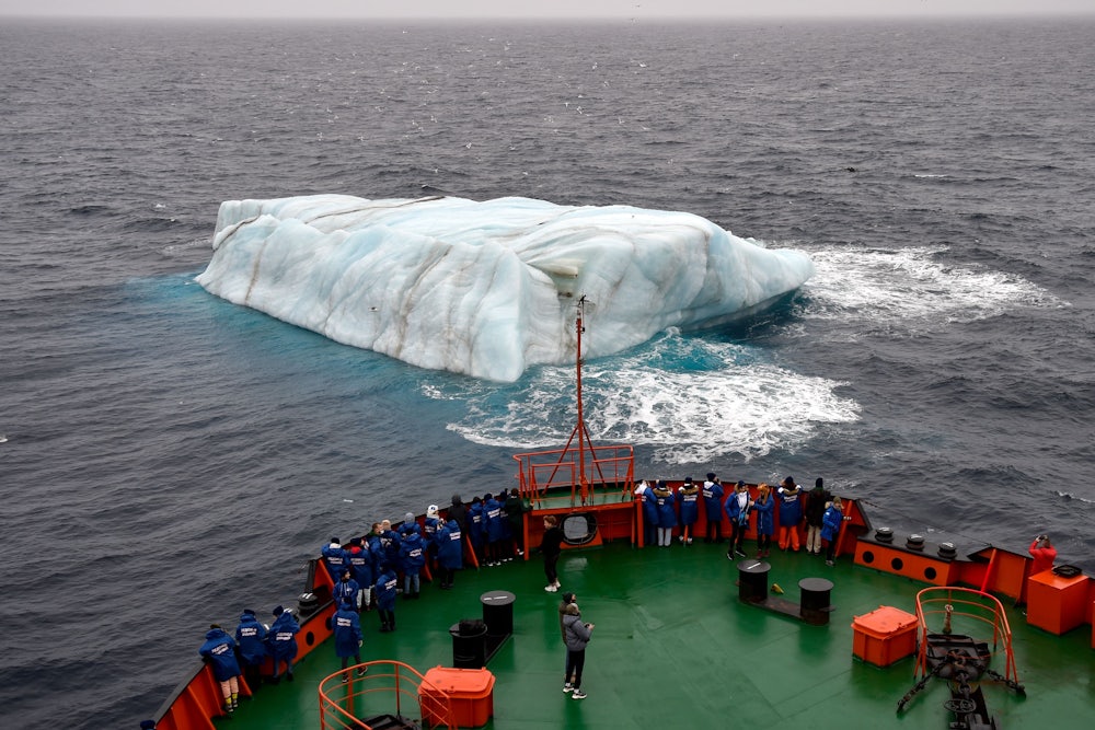People stand on the deck of a ship, looking at an iceberg in the water.
