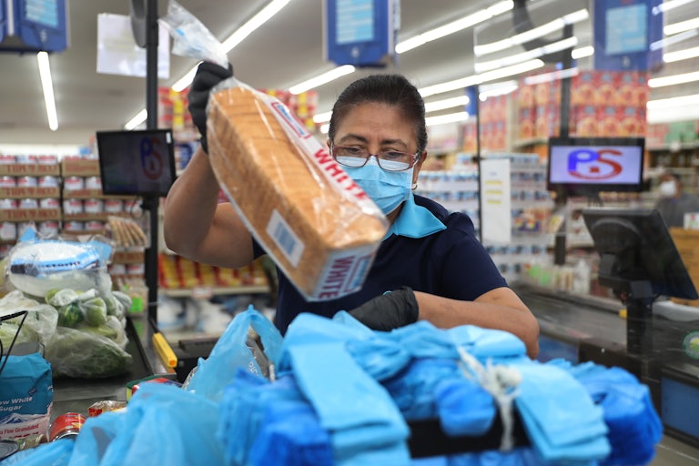 A grocery store worker wearing a mask packs bread into a bag.