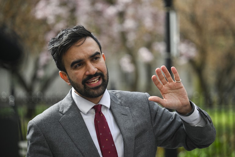 Zohran Mamdani wears a gray suit and holds up his left hand