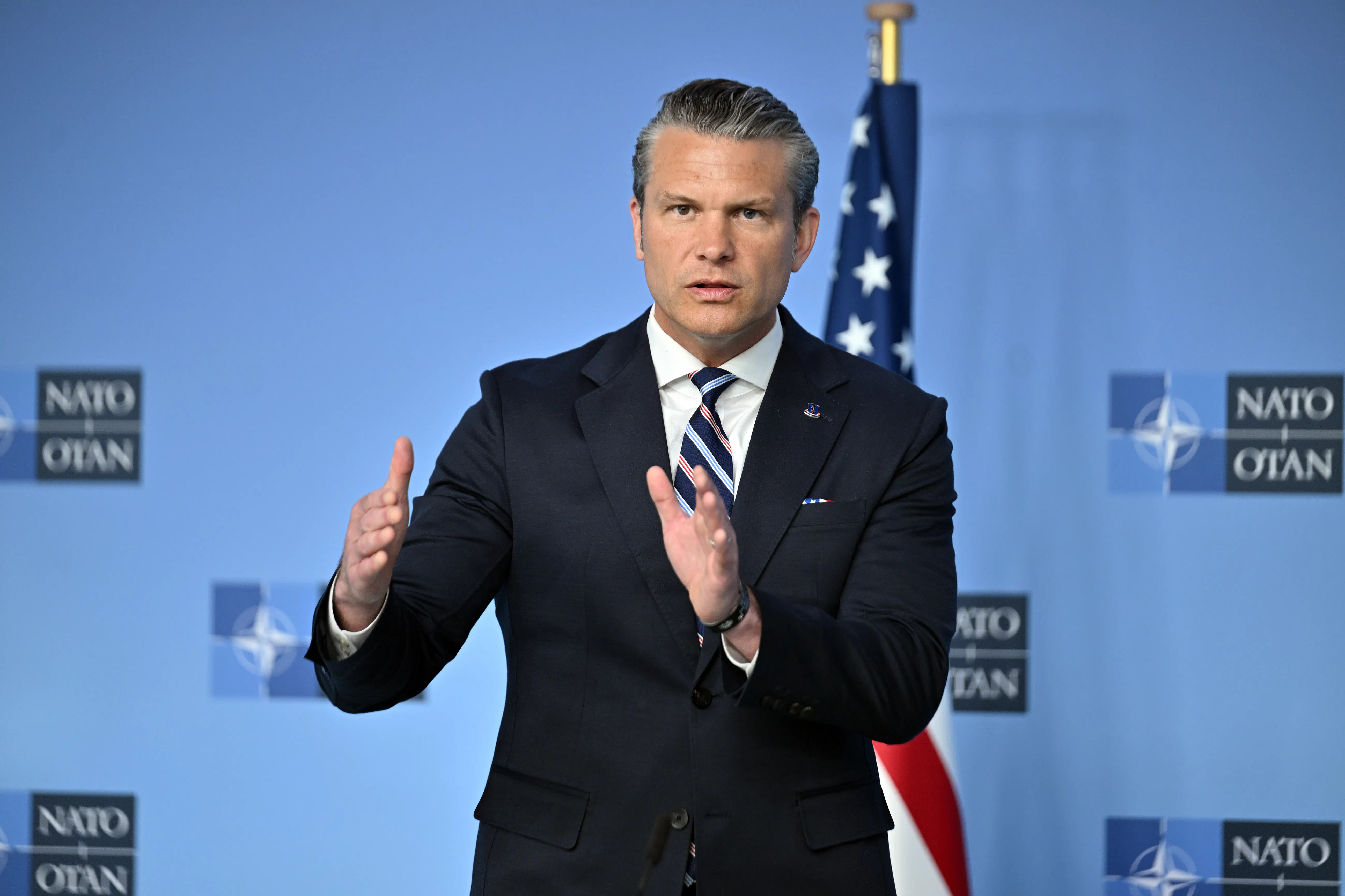 Secretary of Defense Pete Hegseth wearing a suit and striped tie in front of a light blue NATO backdrop with an American flag visible in the background