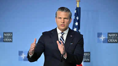 Secretary of Defense Pete Hegseth wearing a suit and striped tie in front of a light blue NATO backdrop with an American flag visible in the background