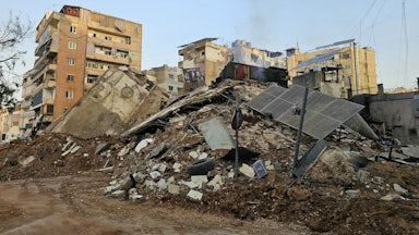 Solar panels lie on a pile of rubble next to other damaged buildings.