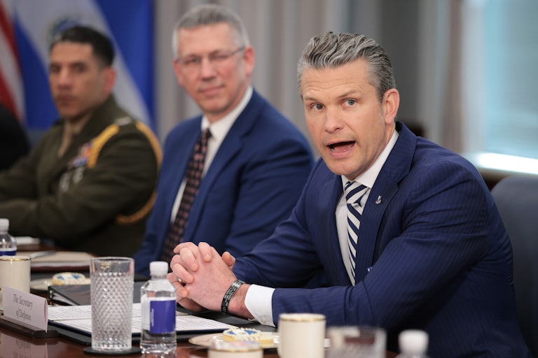 Pete Hegseth has his hands clasped on a table and leans forward slightly as he yells. There are two men seated to his right.