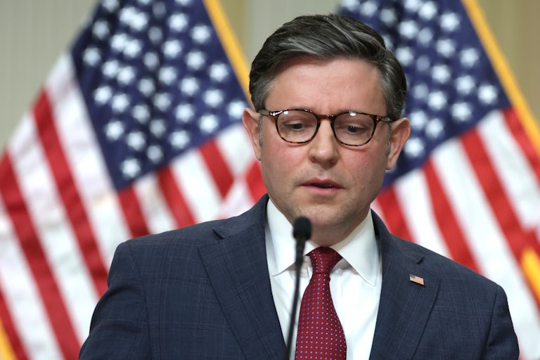 House Speaker Mike Johnson looks down while speaking before a mic. U.S. flags are behind him.