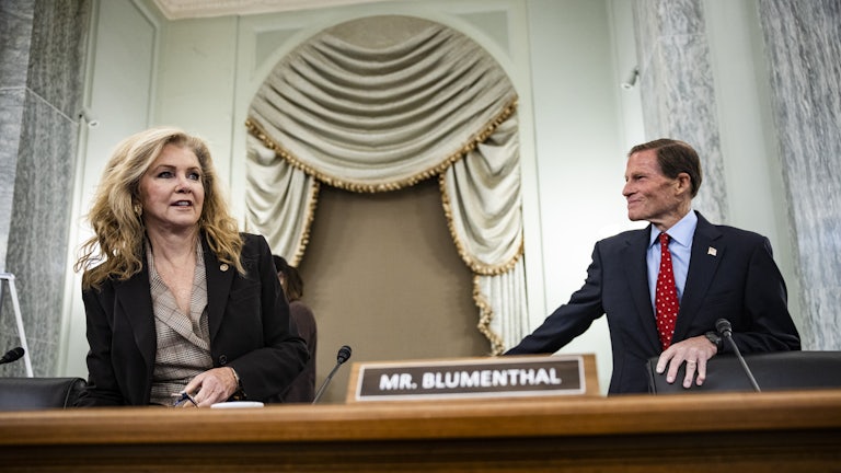Senator Blackburn, a woman with blonde hair, sits down next to Senator Blumenthal, who is standing and looking at her with a smile. Blumenthal's name plaque is pictured between them.