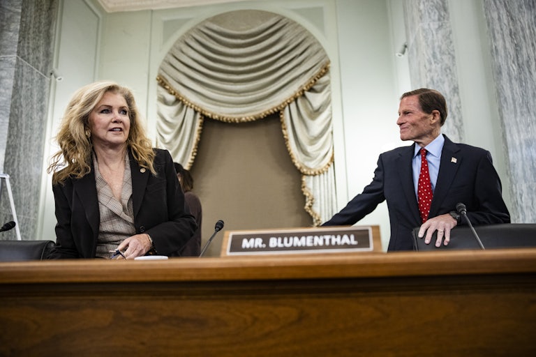 Senator Blackburn, a woman with blonde hair, sits down next to Senator Blumenthal, who is standing and looking at her with a smile. Blumenthal's name plaque is pictured between them.