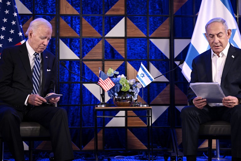 Joe Biden and Benjamin Netanyahu sit in front of U.S. and Israeli flags. Netanyahu is reading from his notes and sitting up straight, while Biden is leaning forward and frowning at his notes sadly. There is a table with a small U.S. and Israeli flag between them.
