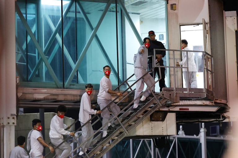 Seven men wearing red face masks, gray sweatpants, and a gray sweatshirt of white T-shirt walk up a set of stairs at the airport. One of them points to the camera.