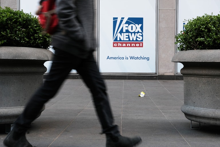 Someone walks by a Fox News sign that has the channel's logo and the line "America is watching." 