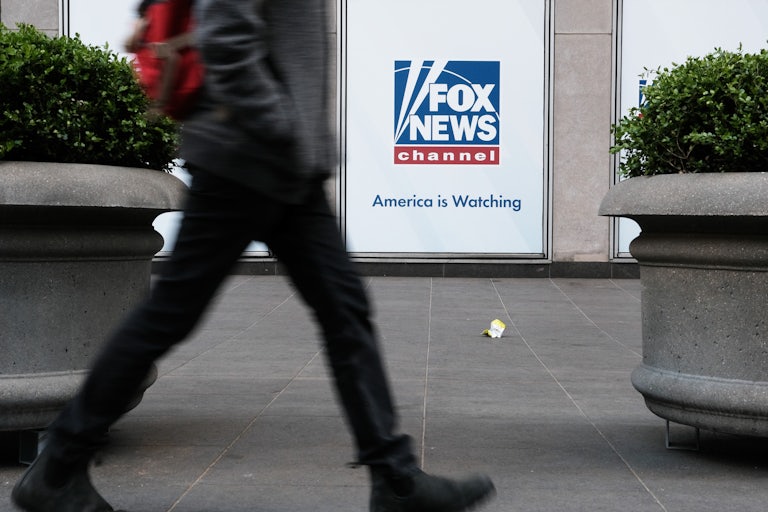 Someone walks by a Fox News sign that has the channel's logo and the line "America is watching."