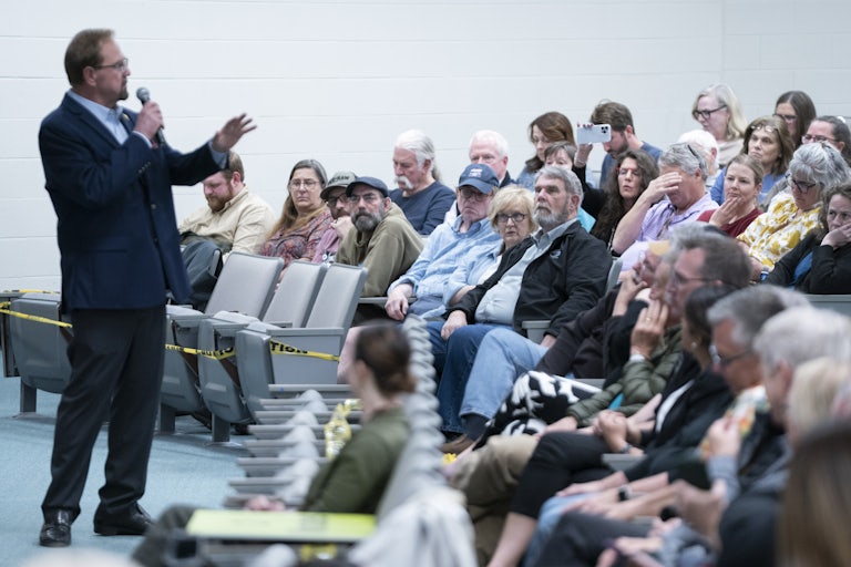 Representative Chuck Edwards speaks into a handheld mic at his town hall. Members of the audience record on their cellphones, listen, or put their head in their hands. One man covers his eyes with his hand as if in disbelief.