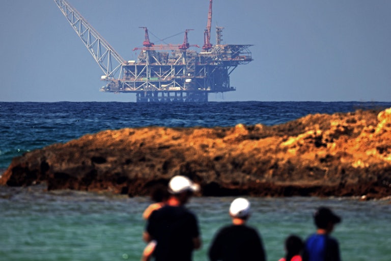 A natural gas rig on the water, seen from shore with people in the foreground 