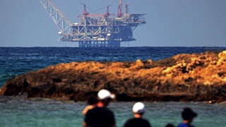 A natural gas rig on the water, seen from shore with people in the foreground