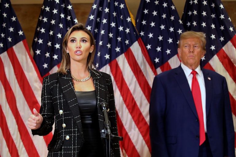 Alina Habba speaks while Donald Trump stands in the background looking at her.