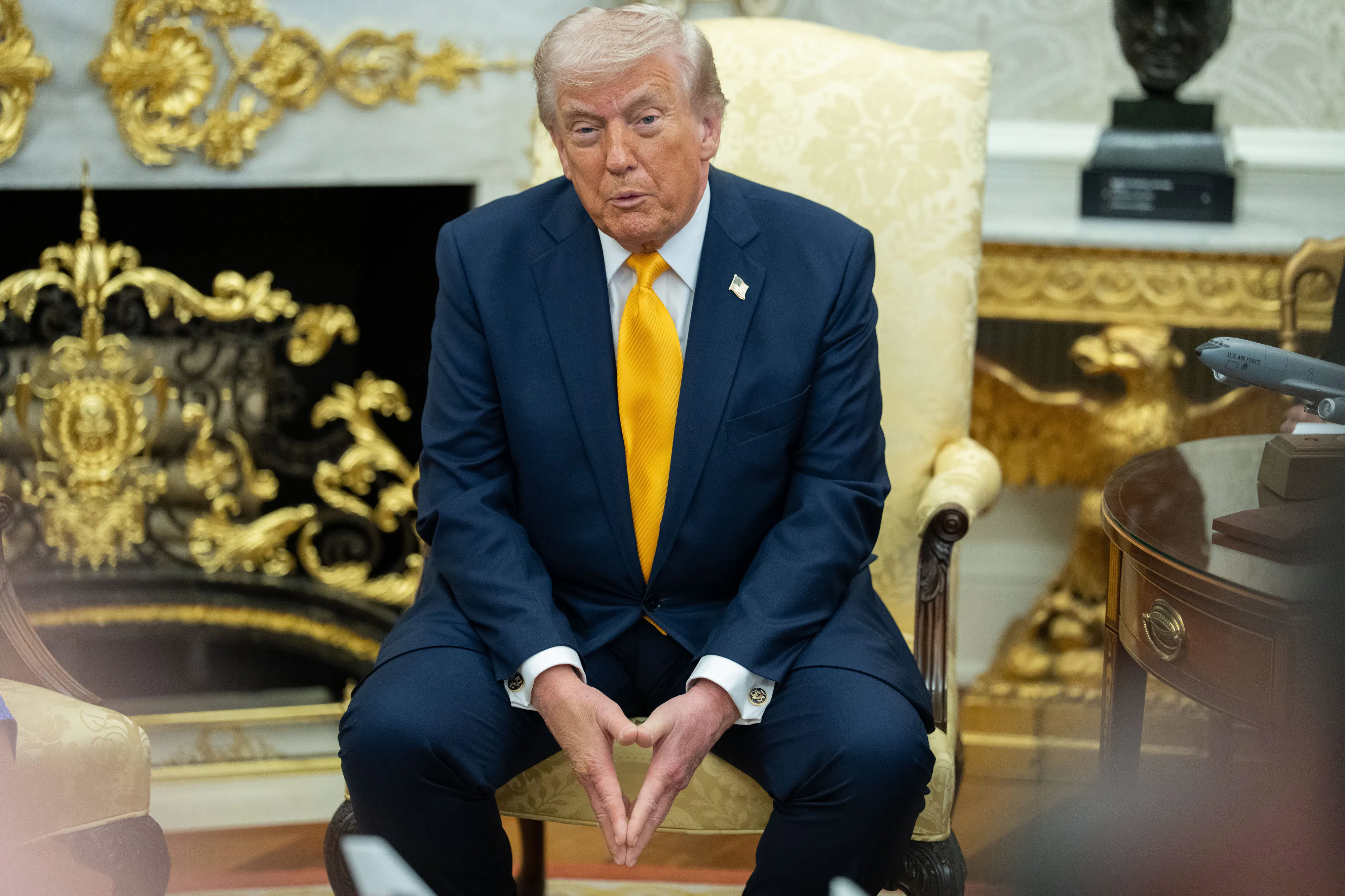 Donald Trump puckers his lips while speaking to reporters. He is seated in an armchair in the Oval Office and has his fingertips pressed together between his legs.