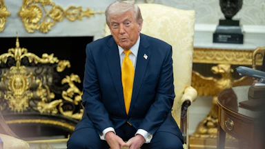 Donald Trump puckers his lips while speaking to reporters. He is seated in an armchair in the Oval Office and has his fingertips pressed together between his legs.