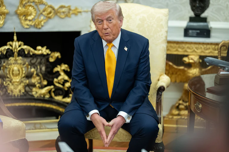 Donald Trump puckers his lips while speaking to reporters. He is seated in an armchair in the Oval Office and has his fingertips pressed together between his legs.