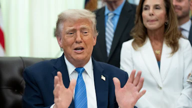 Donald Trump gestures with both hands and speaks while sitting at his desk in the Oval Office
