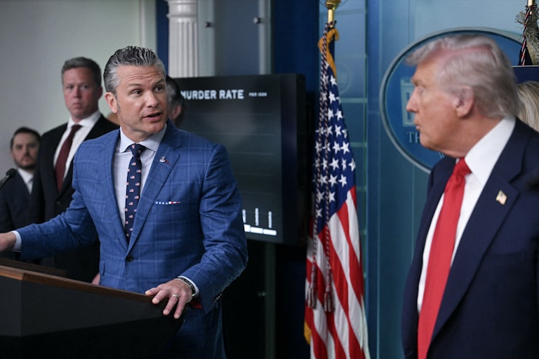 Defense Secretary Pete Hegseth speaks while standing at the lecturn in the White House Press Briefing Room. He turns to look at Donald Trump, who stands nearby.