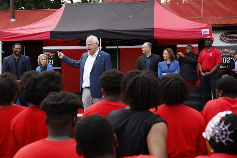Tim Walz speaks in front of the football team players. Kamala Harris and Doug Emhoff smile and look on, standing to his left.