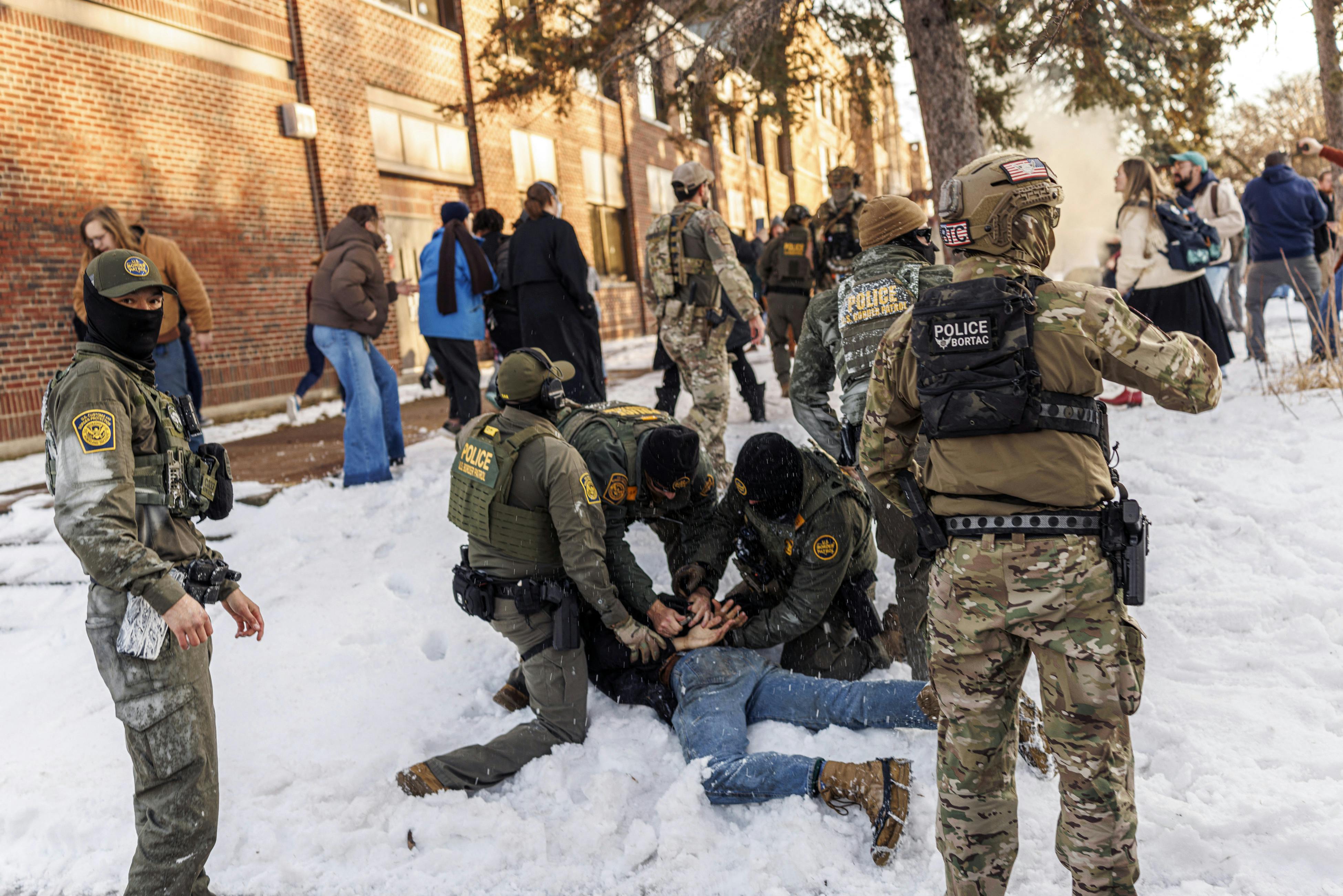 Three masked Border Patrol agents hold a man face-down in the snow, while several other masked agents stand nearby.