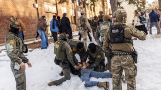 Three masked Border Patrol agents hold a man face-down in the snow, while several other masked agents stand nearby.