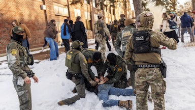 Three masked Border Patrol agents hold a man face-down in the snow, while several other masked agents stand nearby.