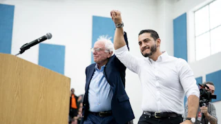 Bernie Sanders and Abdul El-Sayed hold hands in the air in a victorious gesture.