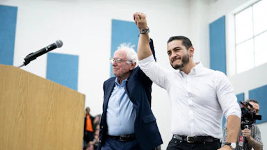 Bernie Sanders and Abdul El-Sayed hold hands in the air in a victorious gesture.