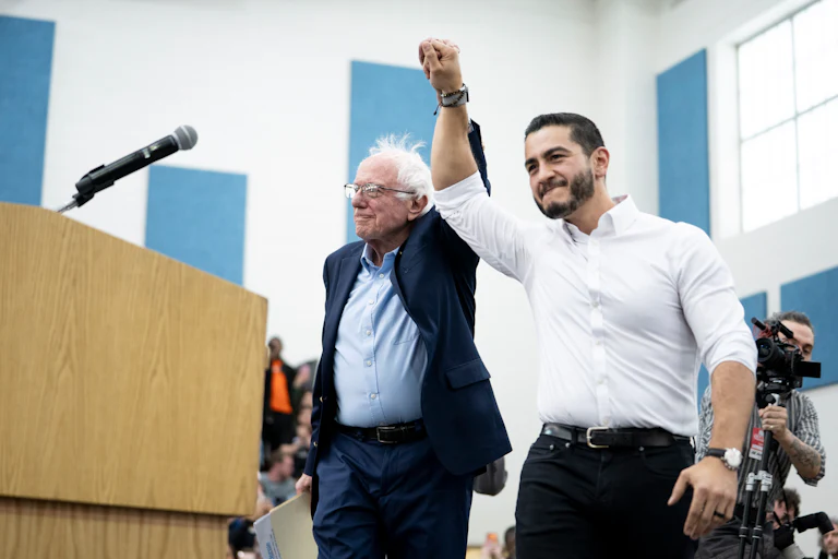 Bernie Sanders and Abdul El-Sayed hold hands in the air in a victorious gesture.