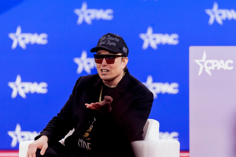 Elon Musk speaks and gestures while sitting on stage at CPAC